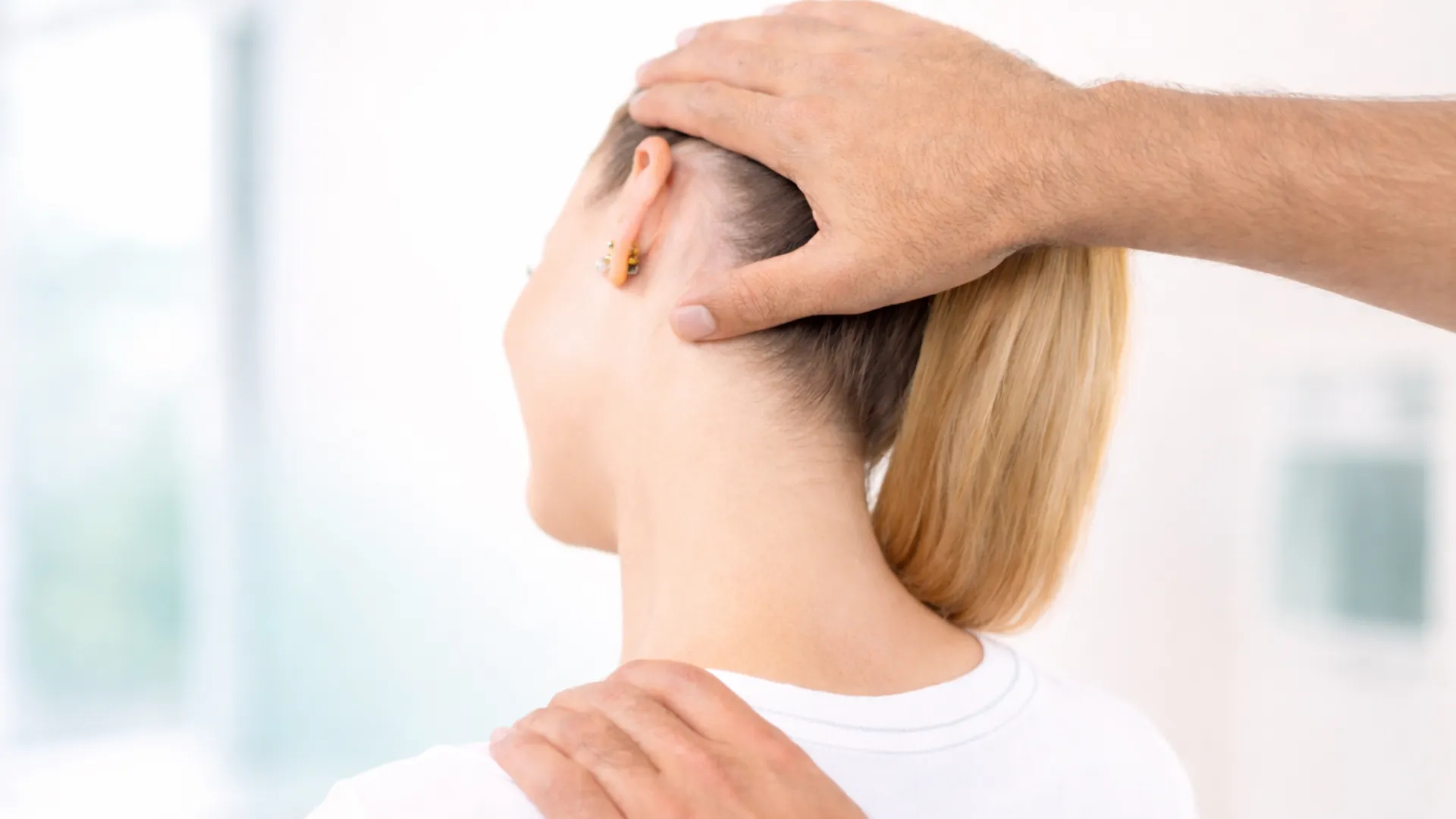 A practitioner’s hands gently support a woman’s head and shoulder as she tilts her neck back, shown in a soft, bright clinical-wellness setting.
